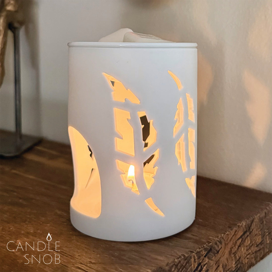 White ceramic lantern with leaf cutouts on a wooden surface, featuring a lit candle inside with reflection on wall.