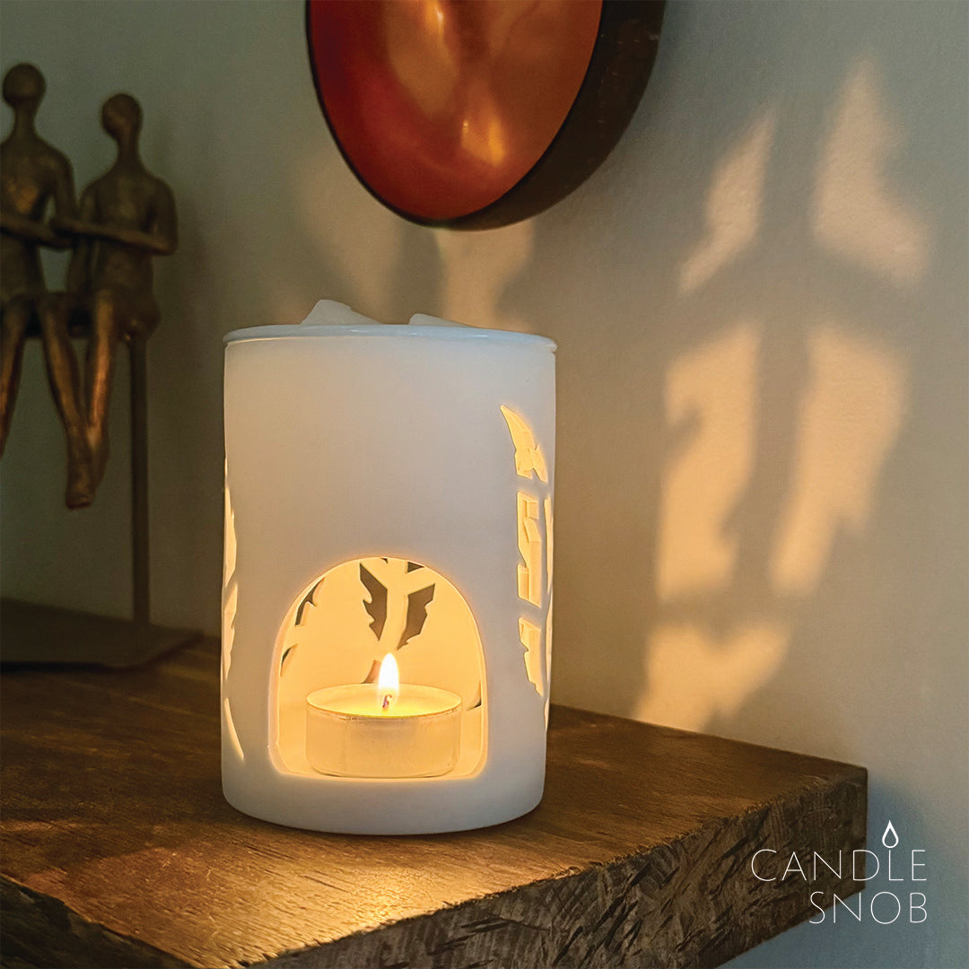 White ceramic feather pattern wax melt burner with a lit candle inside on a wooden shelf, showing reflection on the wall.